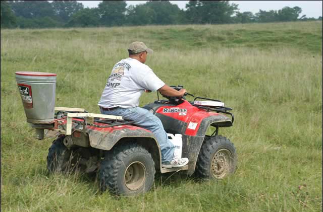 A man riding a four wheeler, in a pasture, with a spreader attached for applying granular fire ant bait.