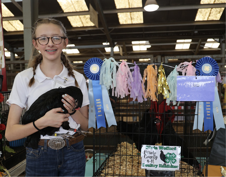 A young girl in glasses holds her bird while standing in front of a cage decorated with blue prize ribbons. 