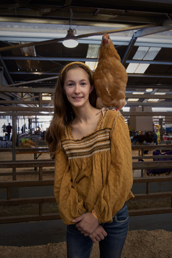 A young girl stands with a bird on her left shoulder. Her brownish shirt and hair band match the color of the bird.