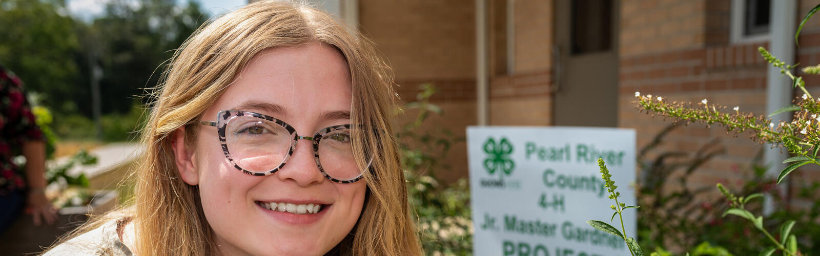 A person smiling, holding a plant in front of a sign listing, “Pearl River County 4-H Jr. Master Gardener Project.”