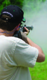 A young man wearing a white shirt and black cap, aims and shoots a muzzleloader causing smoke to fill the air.